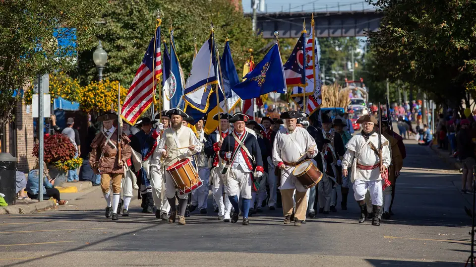 Colonial reenactors with flags and drums marching down Main Street during a festival parade